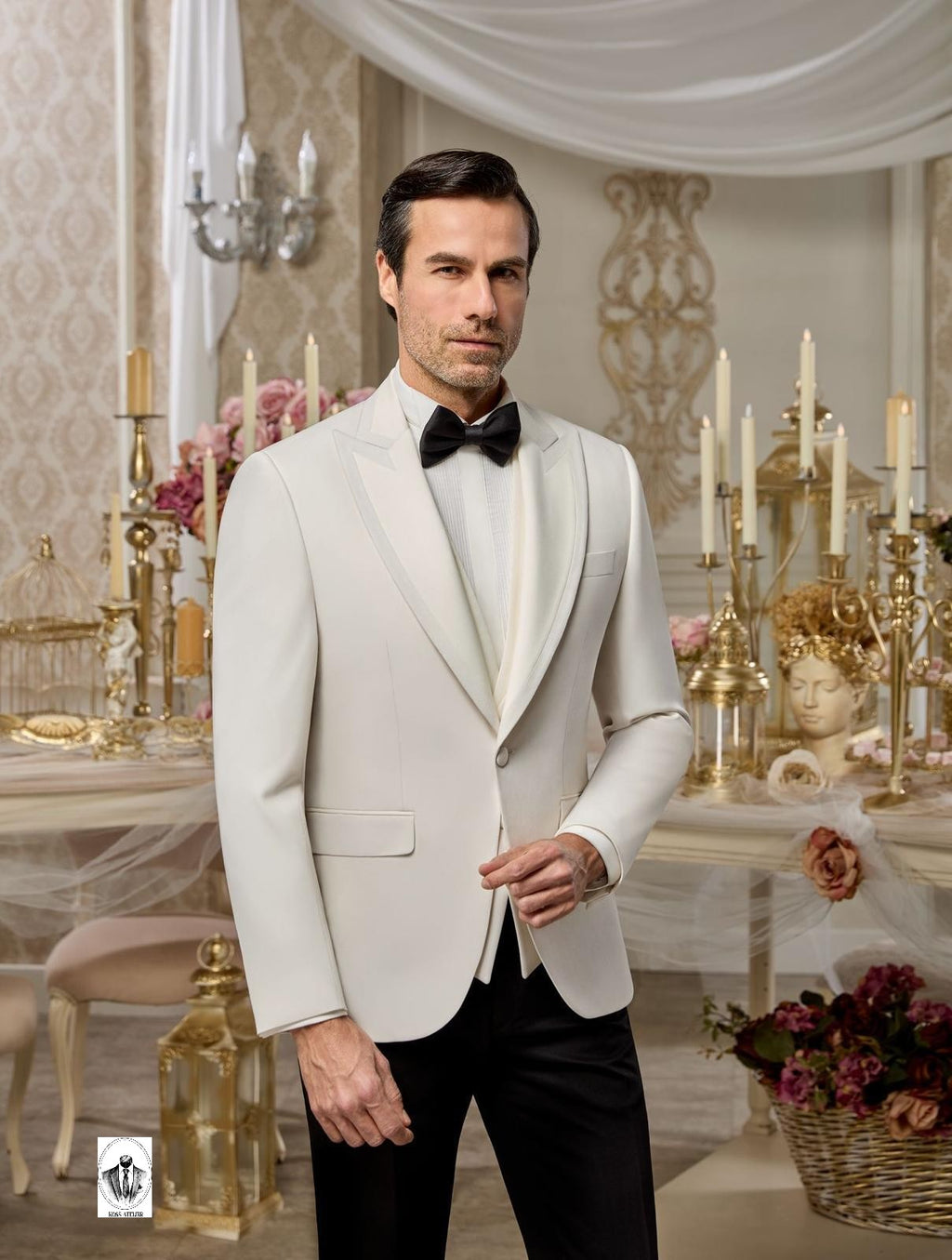 A man wearing a light ivory or off-white tuxedo jacket with satin peak lapels, paired with a white shirt, black bow tie, and black trousers, standing in an ornate, elegant room decorated with candles, flowers, and classical statues.