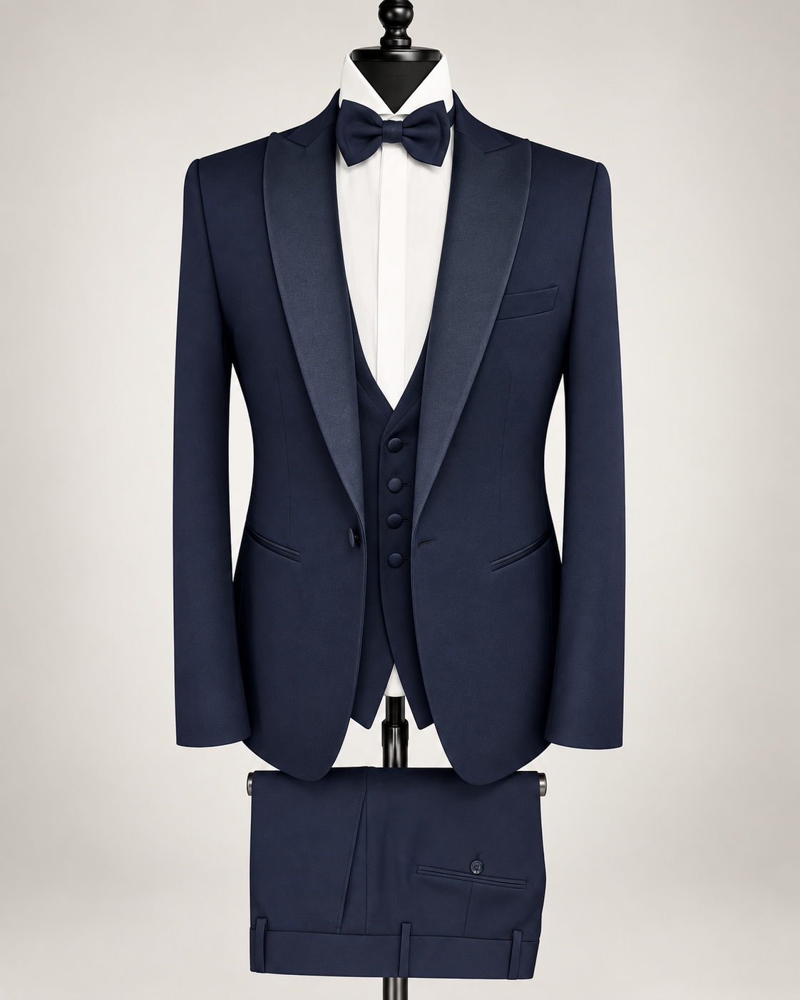 A navy blue formal tuxedo set displayed on a black mannequin against a light gray background. The set includes a single-breasted jacket with satin peak lapels, a matching waistcoat, trousers folded below, a white dress shirt, and a matching navy bow tie.