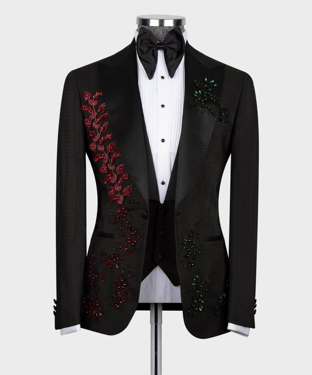 A black tuxedo jacket with red and green sequined floral embroidery on the lapels, styled on a mannequin with a white pinstripe shirt, black bow tie, and black vest against a light gray background.
