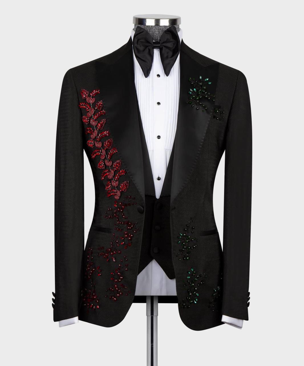 A black tuxedo jacket with red and green sequined floral embroidery on the lapels, styled on a mannequin with a white pinstripe shirt, black bow tie, and black vest against a light gray background.
