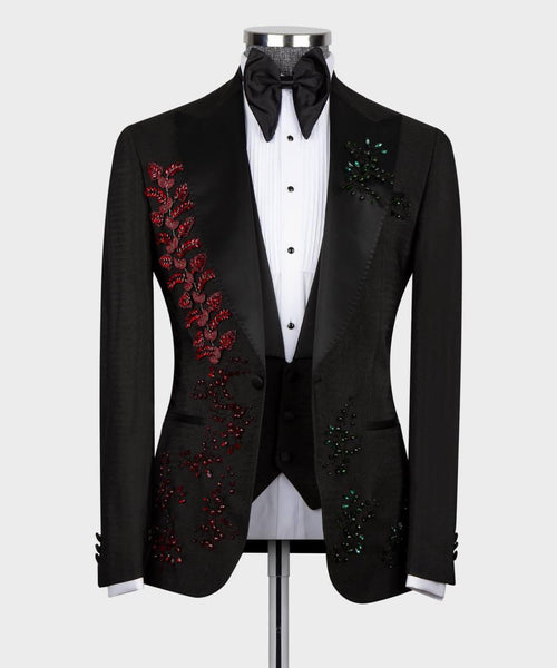 A black tuxedo jacket with red and green sequined floral embroidery on the lapels, styled on a mannequin with a white pinstripe shirt, black bow tie, and black vest against a light gray background.