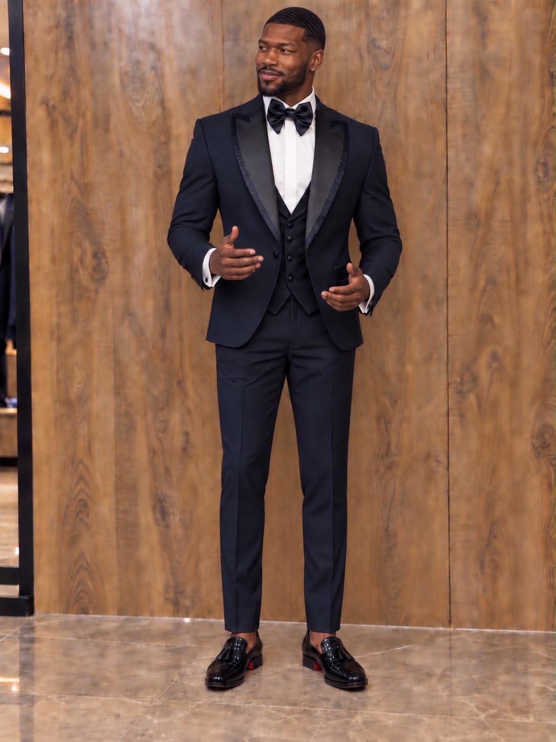 A man wearing a black tuxedo with black satin lapels, a white dress shirt, a black bow tie, and a black vest, standing in front of a wood-paneled wall.