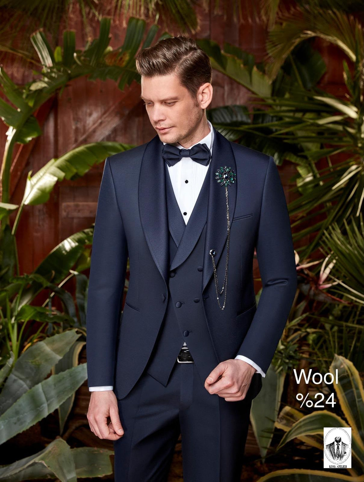 A man wearing a dark navy blue three-piece tuxedo with a white dress shirt, black bow tie, and a decorative brooch, posed in front of a tropical background with green leaves and a wooden wall. Text on the bottom right reads 'Wool %24' with a small logo.