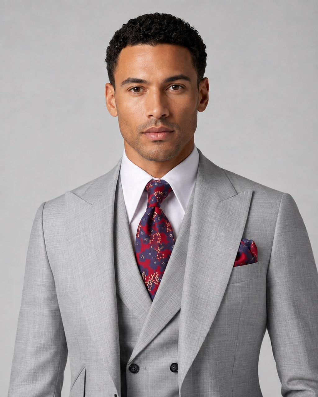 A man wearing a light gray three-piece suit with a white dress shirt, a red and blue patterned tie, and a matching pocket square, photographed against a neutral gray background.
