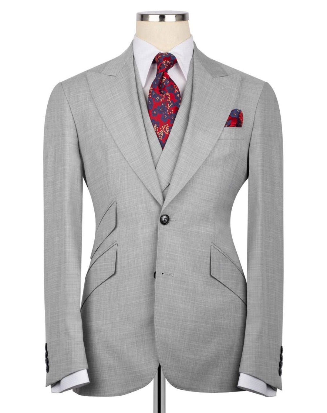 A light gray three-piece suit displayed on a mannequin, styled with a white dress shirt, a red and blue paisley patterned tie, and a matching pocket square.