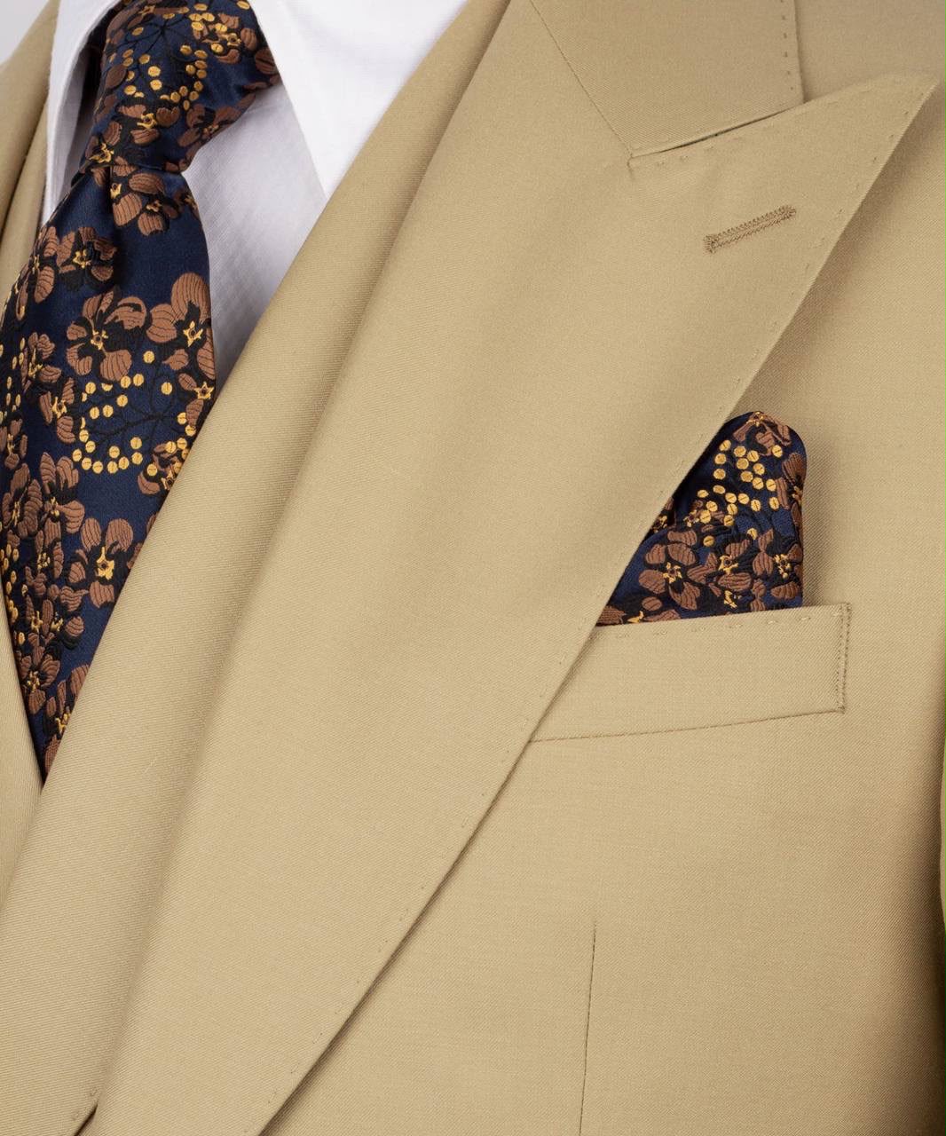 A close-up of a beige suit jacket with a matching pocket square and a navy blue patterned tie with gold floral details, worn with a white dress shirt.