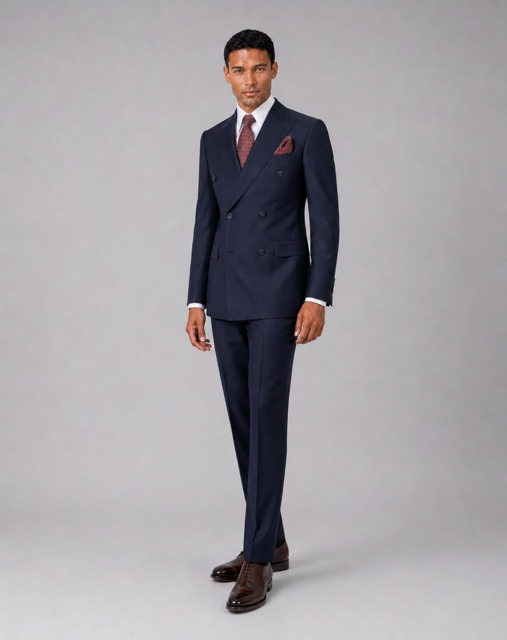 A man modeling a navy blue double-breasted suit with a white dress shirt, a burgundy patterned tie, a matching pocket square, and dark brown dress shoes, standing against a solid gray studio background.
