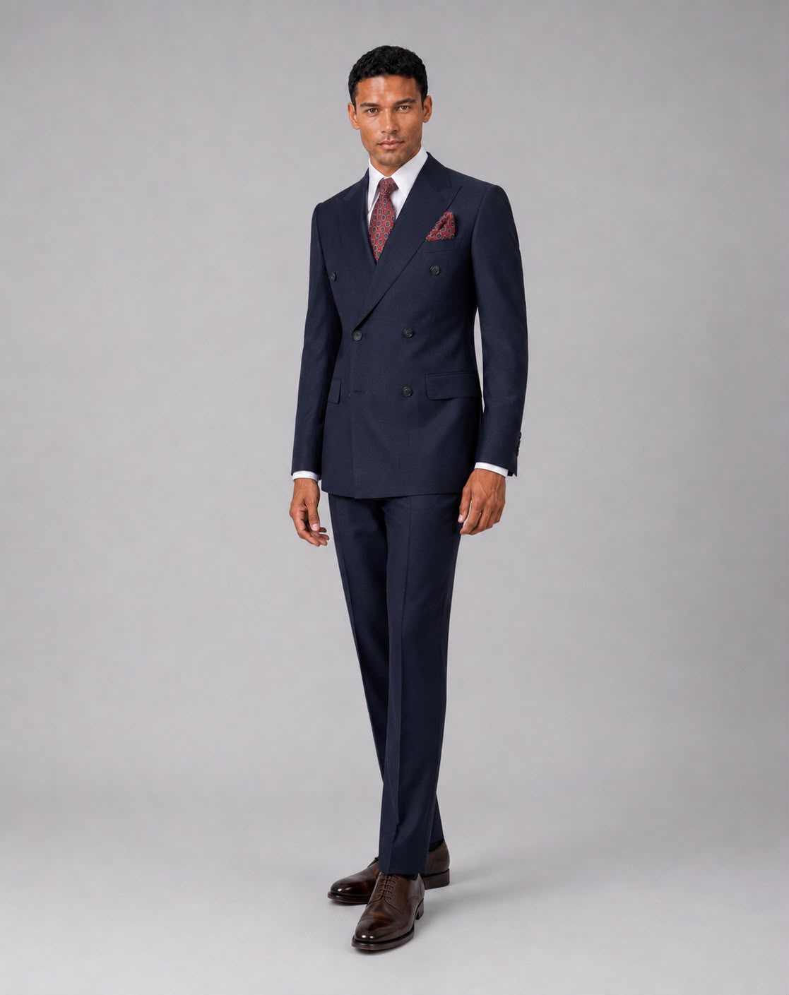A man modeling a navy blue double-breasted suit with a white dress shirt, a burgundy patterned tie, a matching pocket square, and dark brown dress shoes, standing against a solid gray studio background.