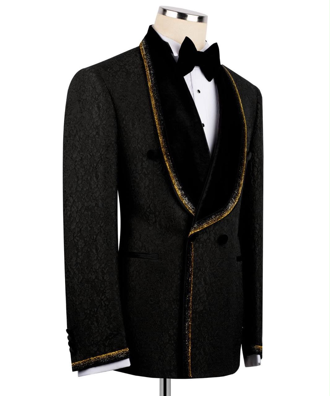 A black double-breasted tuxedo jacket with a textured pattern, black velvet shawl lapels, and gold glitter trim, styled on a mannequin with a white shirt and black bow tie.