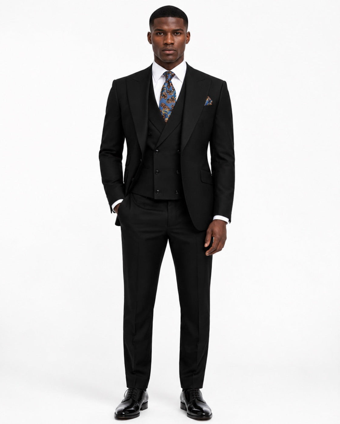 A man modeling a black three-piece suit with a white dress shirt, a patterned blue and gold tie, and a matching pocket square.