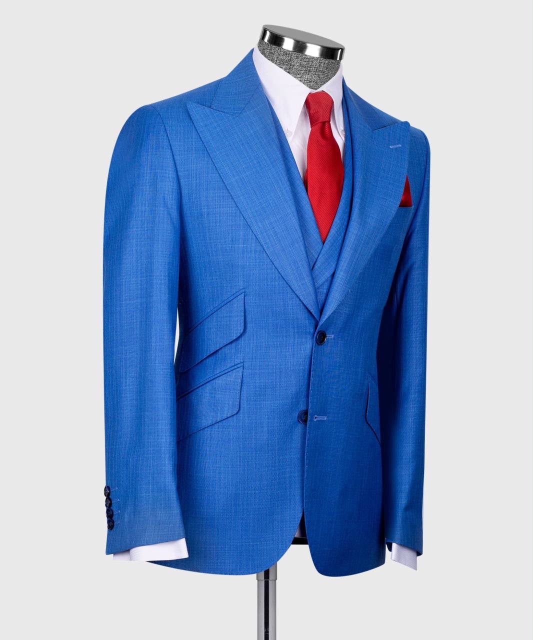 A bright blue two-piece suit with a subtle micro-check pattern, displayed on a mannequin torso with a white dress shirt, red tie, and matching red pocket square against a light gray background.