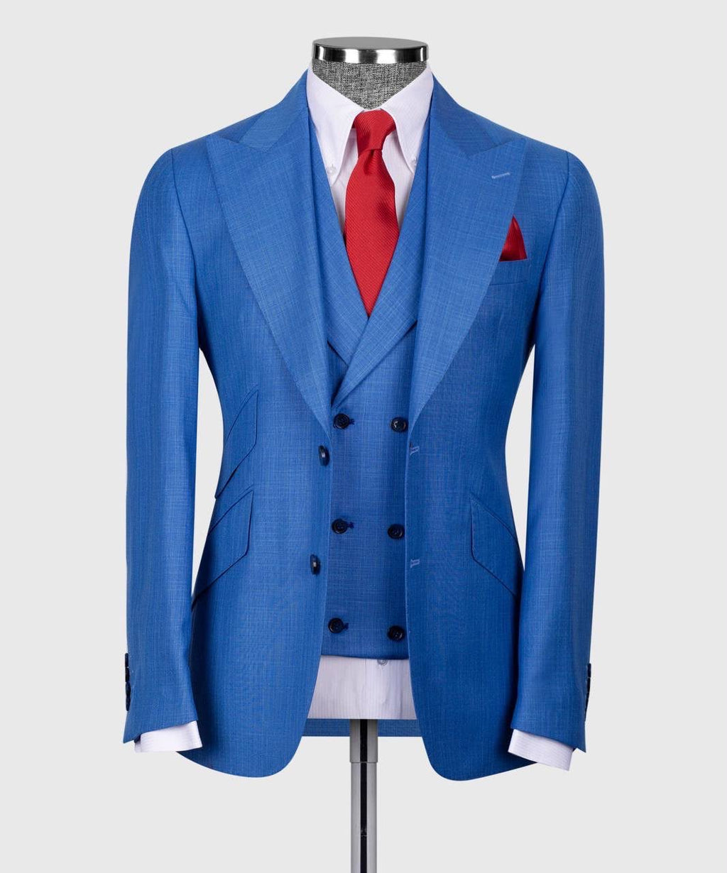 A men's three-piece suit displayed on a mannequin, featuring a blue double-breasted blazer, matching blue vest, and a blue patterned tie, styled with a white dress shirt and a red pocket square.