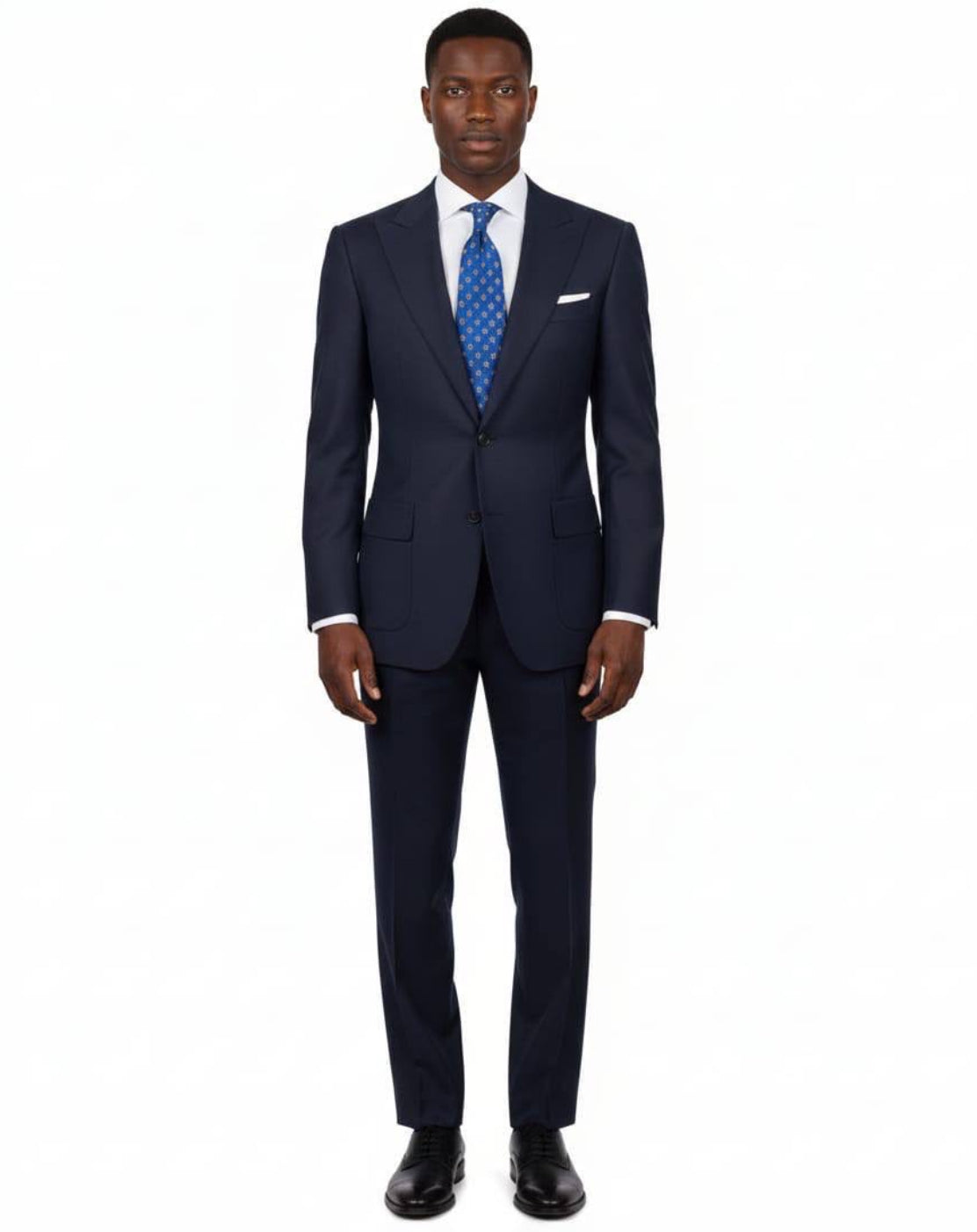 A man modeling a navy blue two-piece suit with a white dress shirt, a blue patterned tie, and a white pocket square, standing against a plain white background.