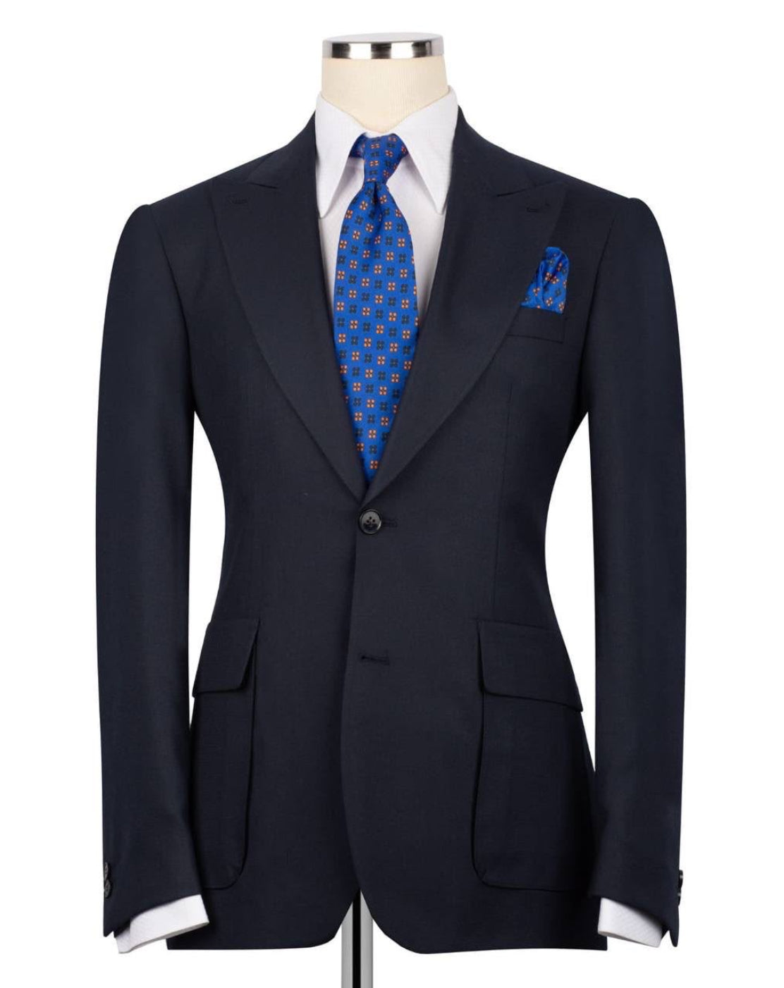 A dark navy blue suit jacket displayed on a mannequin torso, styled with a white dress shirt, a blue patterned tie, and a matching blue patterned pocket square against a plain white background.