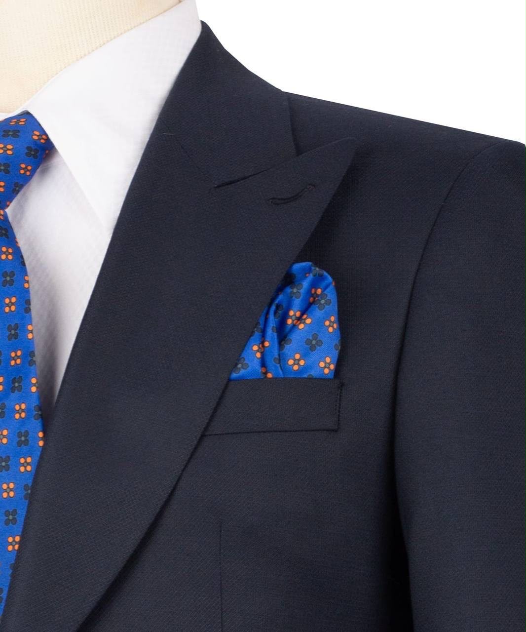 A close-up of a dark navy suit jacket on a mannequin, styled with a white dress shirt, a blue patterned tie, and a matching blue patterned pocket square.