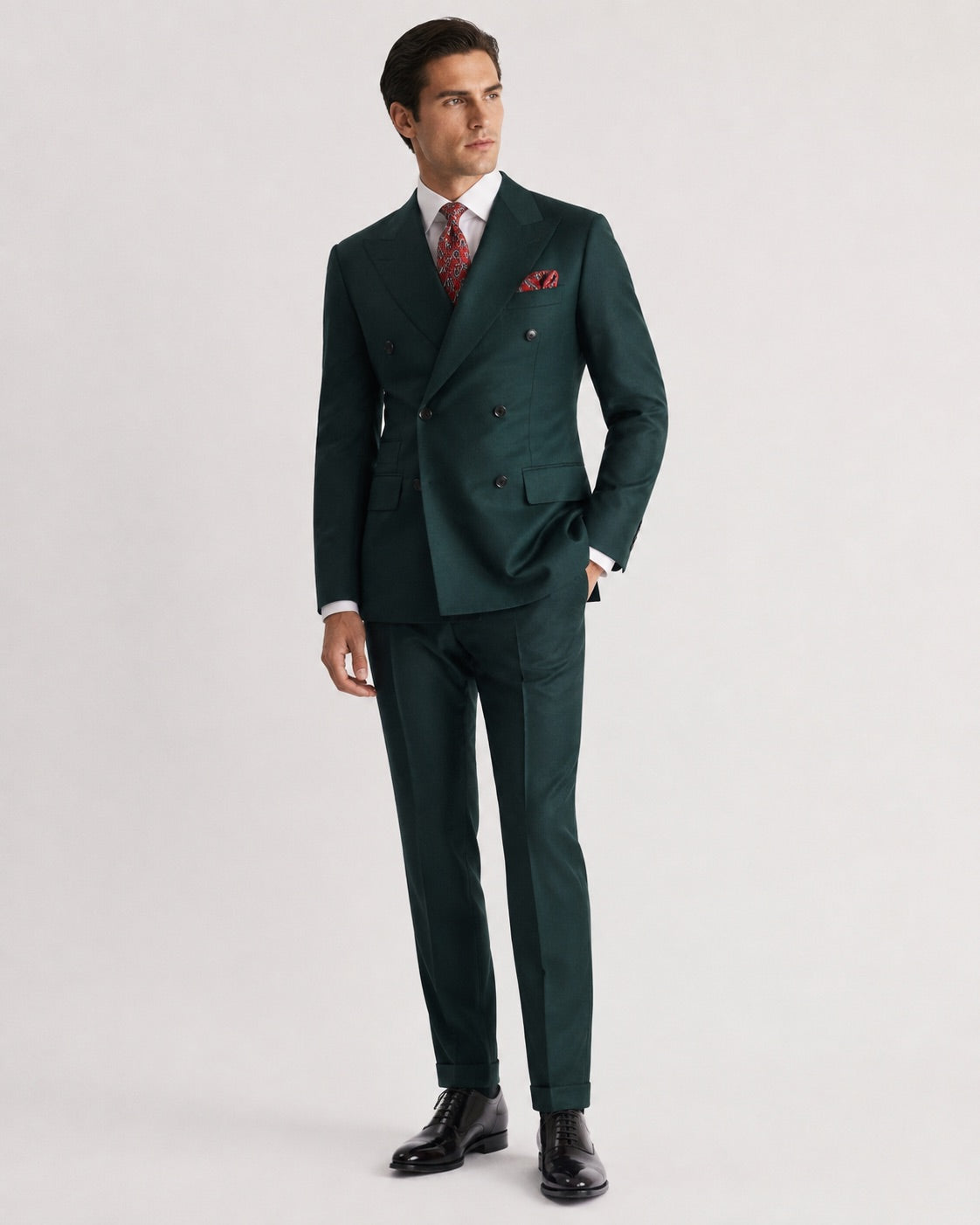 A man modeling a dark green double-breasted suit with a white dress shirt, a red patterned tie, and a matching red pocket square