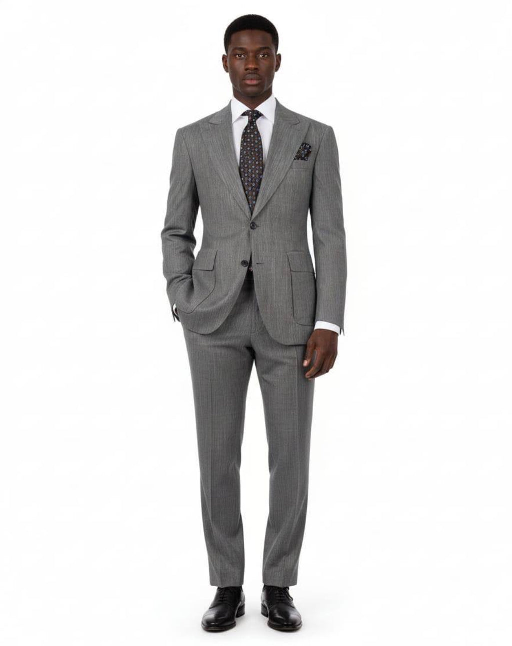 A man modeling a gray two-piece suit with a subtle micro-check pattern, paired with a white dress shirt, a patterned dark tie, and black dress shoes, against a plain white background.