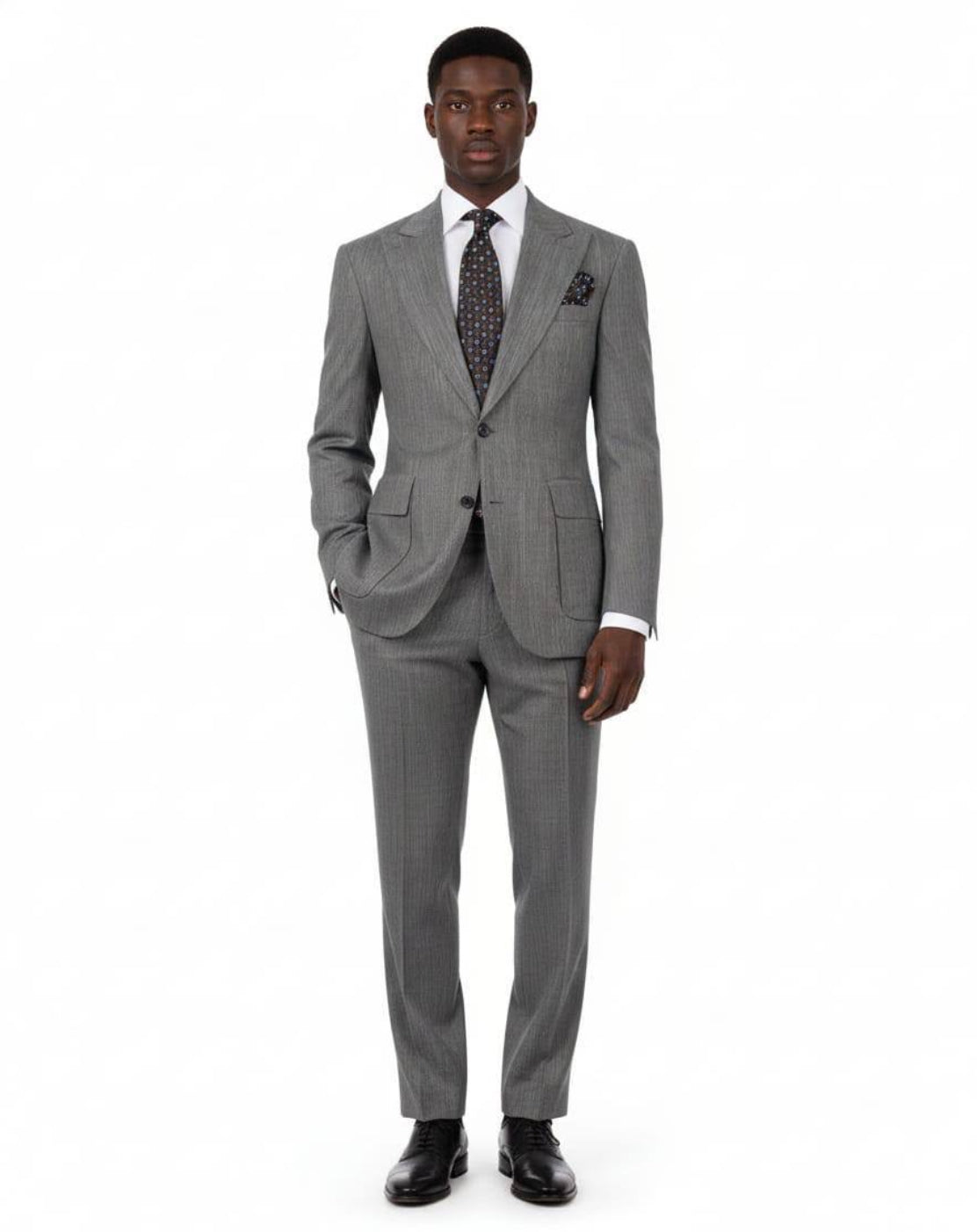 A man modeling a gray two-piece suit with a subtle micro-check pattern, paired with a white dress shirt, a patterned dark tie, and black dress shoes, against a plain white background.