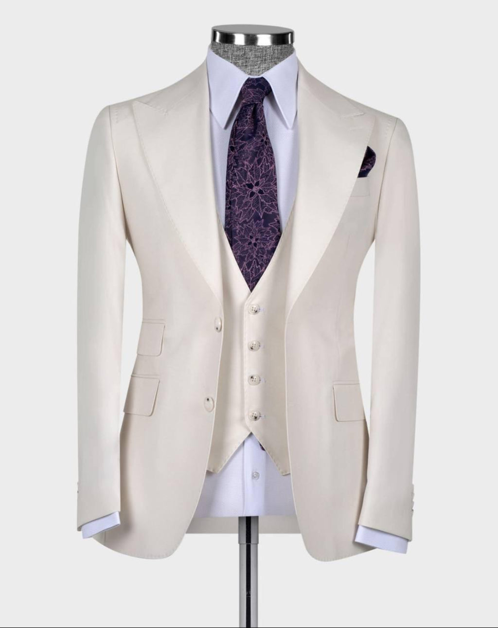 A cream-colored three-piece suit displayed on a mannequin, featuring a tailored jacket, matching vest, white dress shirt, a purple patterned tie, and a matching pocket square against a light gray background.