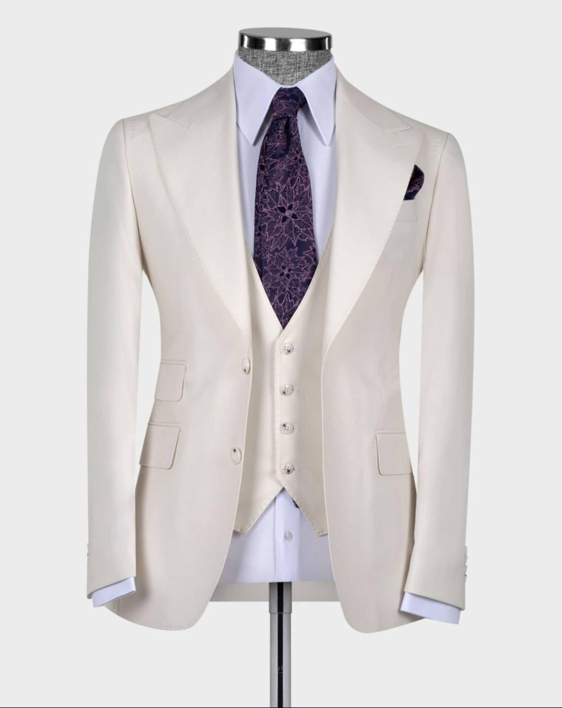 A cream-colored three-piece suit displayed on a mannequin, featuring a tailored jacket, matching vest, white dress shirt, a purple patterned tie, and a matching pocket square against a light gray background.