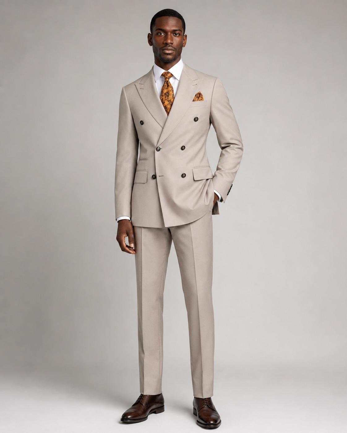 A man modeling a light beige double-breasted suit with a white dress shirt, an orange patterned tie, and a matching pocket square, standing against a neutral gray studio background.
