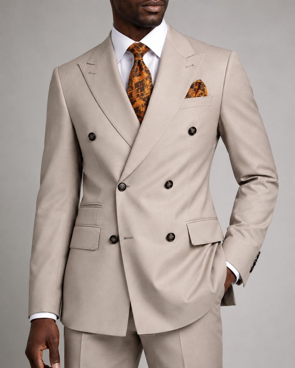 A man wearing a light beige double-breasted suit with a white dress shirt, an orange patterned tie, and a matching orange pocket square, photographed against a neutral gray studio background.