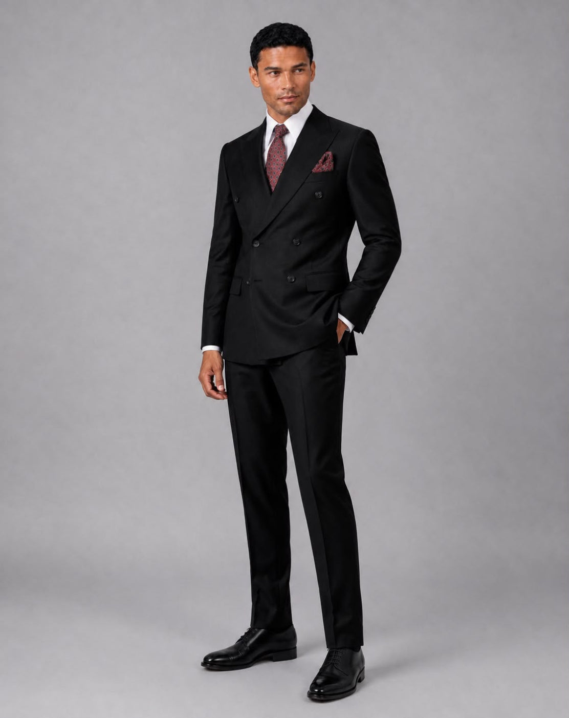 A man modeling a black double-breasted suit with a white dress shirt, a patterned burgundy tie, and a matching pocket square against a solid gray studio background.
