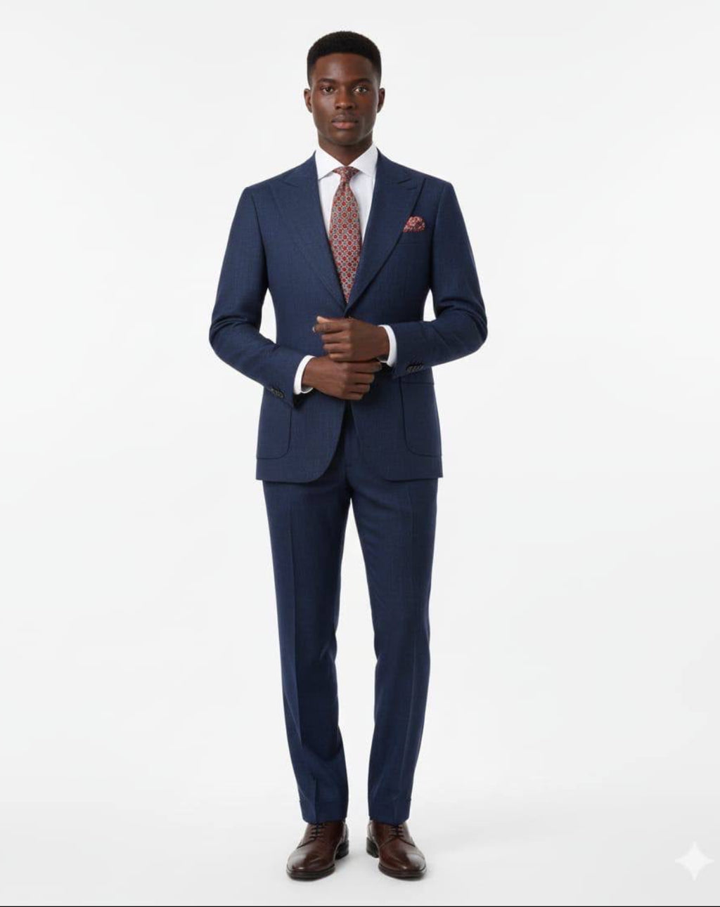 A man modeling a navy blue two-piece suit with a white dress shirt, a patterned reddish-brown tie, a matching pocket square, and brown dress shoes, standing against a plain white studio background.