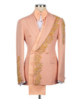 Men's luxury double-breasted tuxedo jackets shown in midnight black and pastel peach, featuring an innovative asymmetrical extended sash and heavy gold crystal floral appliqués on the lapel, shoulder, and cuffs