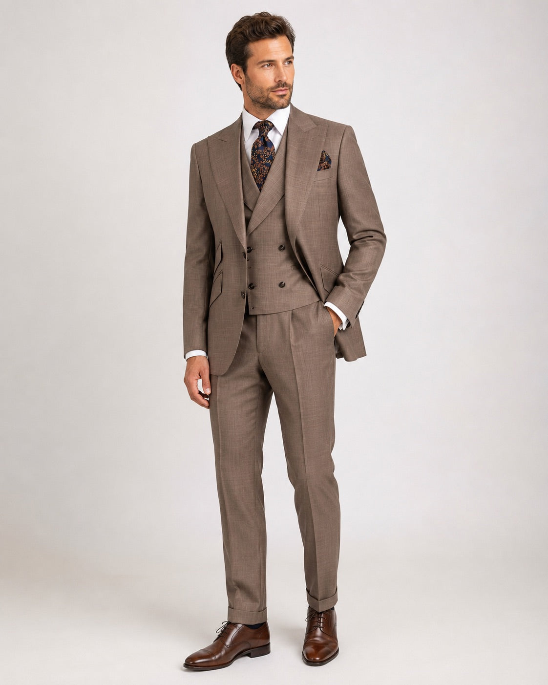 A man modeling a brown three-piece suit with a subtle pinstripe pattern, featuring a double-breasted jacket, matching vest, and trousers, styled with a white shirt, patterned tie, and brown leather dress shoes 