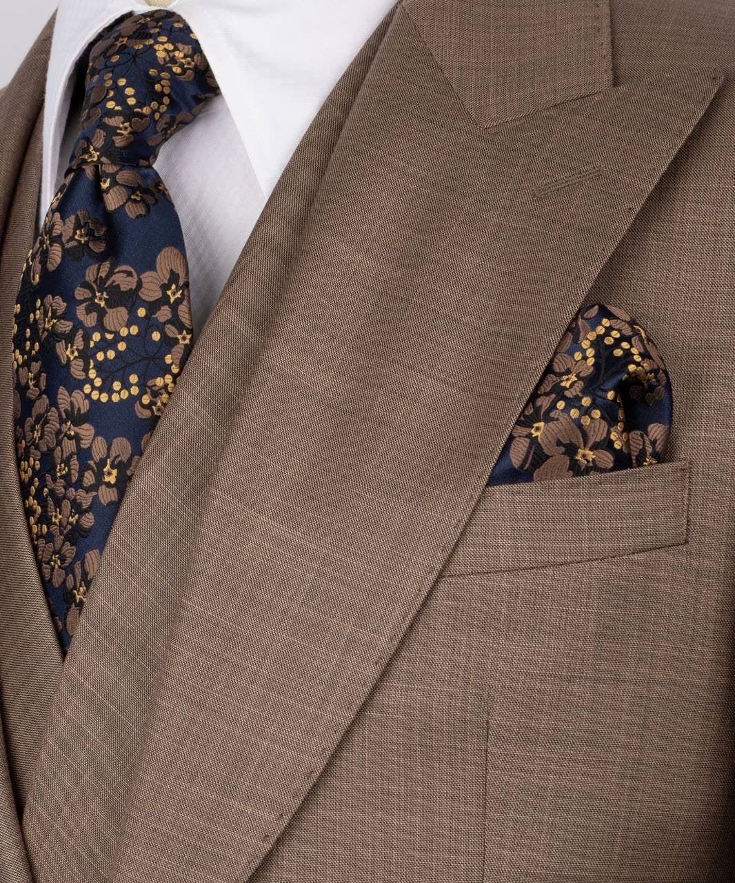 A close-up of a men's formal suit featuring a brown and beige textured wool jacket, a navy blue floral silk tie, and a matching pocket square.