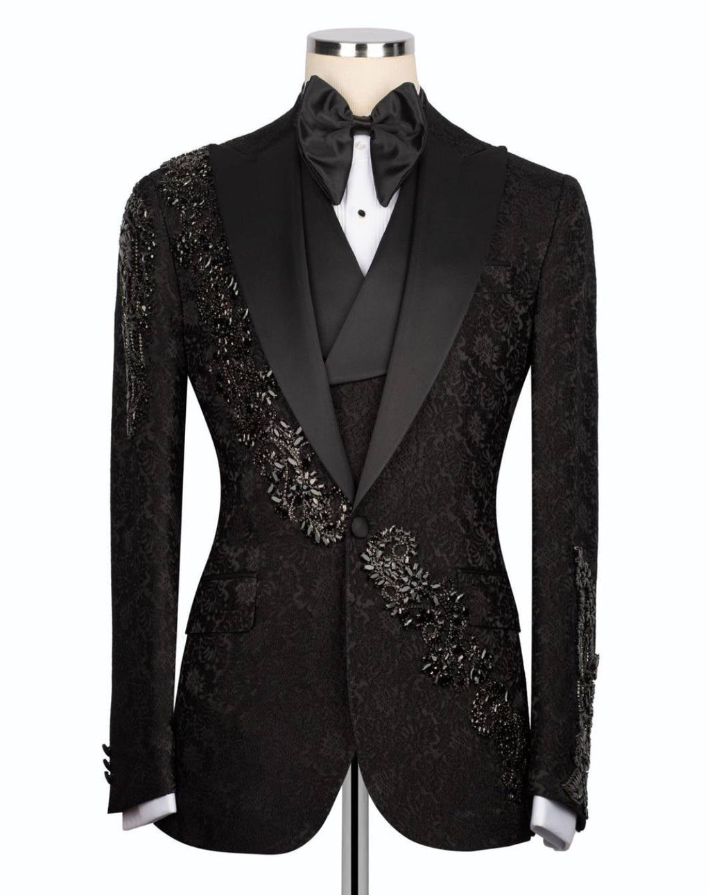 A black tuxedo jacket with ornate beaded and embroidered detailing on the shoulders and chest, styled on a mannequin with a black satin bow tie and white shirt.