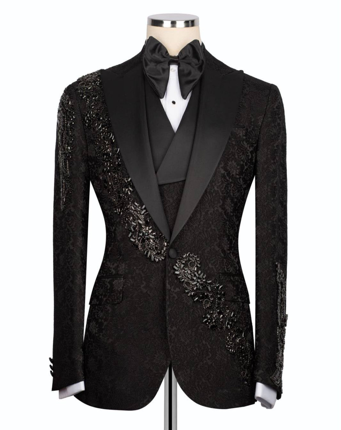 A black tuxedo jacket with ornate beaded and embroidered detailing on the shoulders and chest, styled on a mannequin with a black satin bow tie and white shirt.