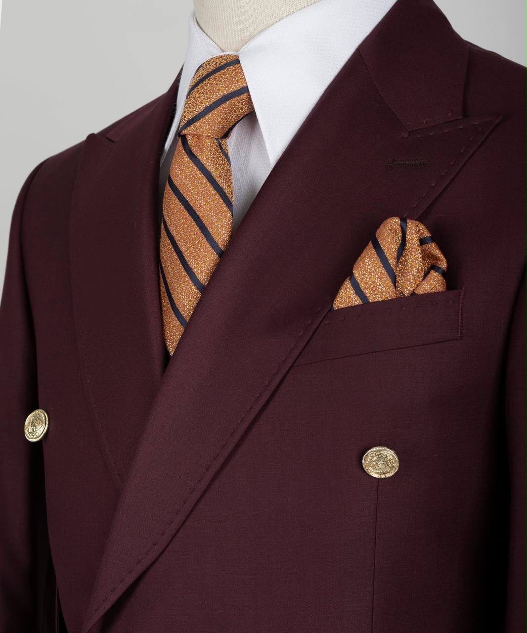 A close-up of a double-breasted suit jacket in deep burgundy, styled with a white dress shirt, a gold-striped tie, and a matching pocket square.