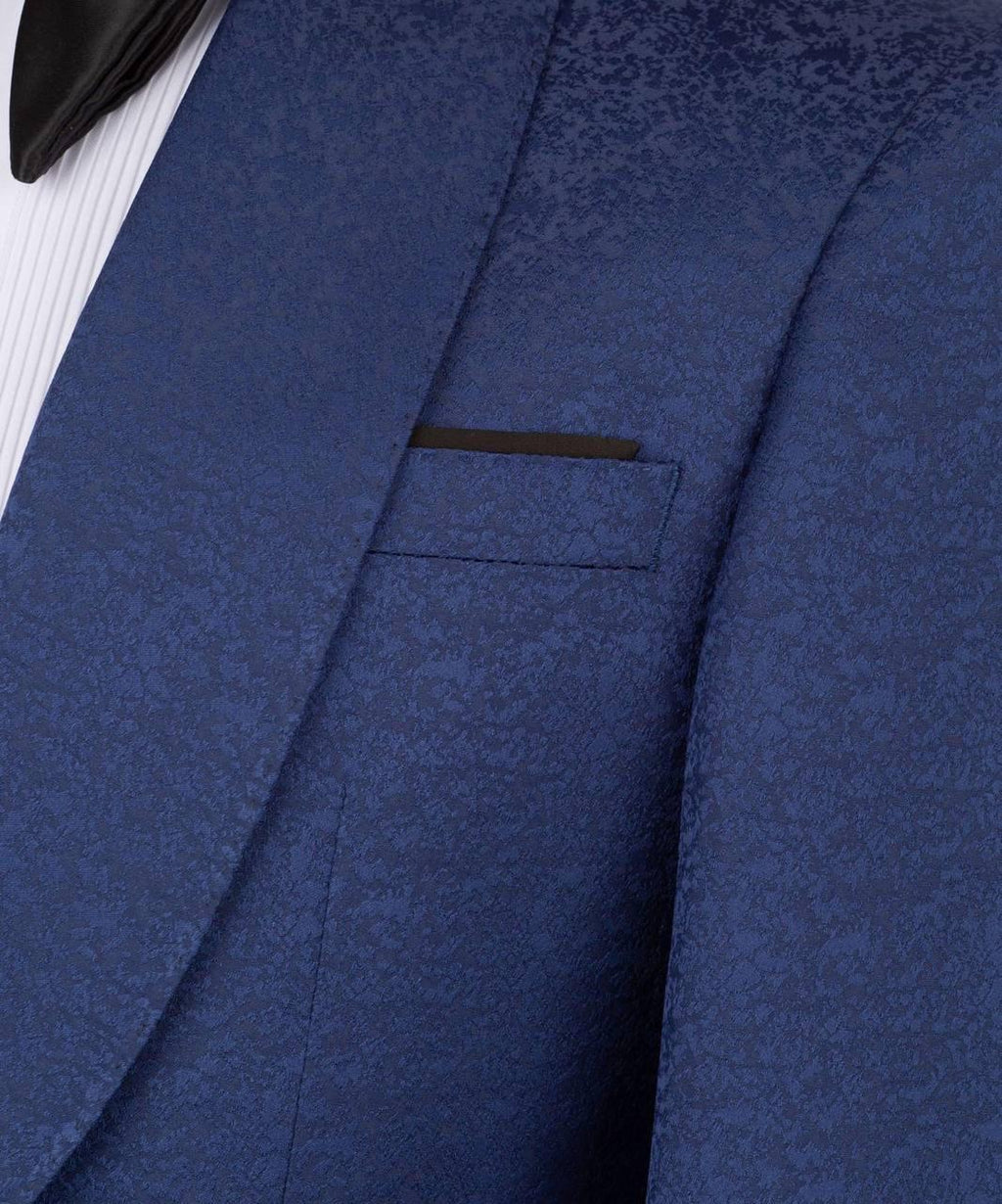 Close-up of a textured blue patterned suit jacket with a black pocket square and a white pleated shirt visible at the edge.