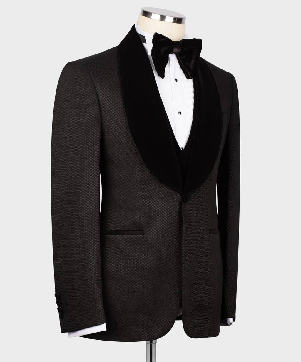 A black tuxedo jacket with a wide black velvet shawl collar, displayed on a mannequin torso with a white dress shirt, black bow tie, and black vest against a light gray background.
