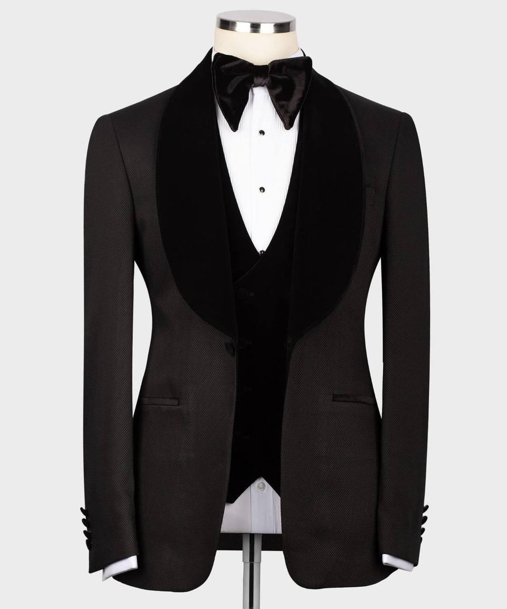 A black tuxedo jacket with wide black velvet shawl lapels, displayed on a mannequin torso with a black bow tie, black vest, and white dress shirt against a light gray background.
