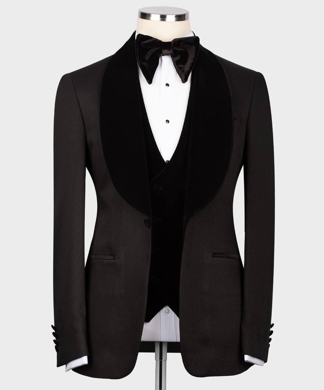 A black tuxedo jacket with wide black velvet shawl lapels, displayed on a mannequin torso with a black bow tie, black vest, and white dress shirt against a light gray background.