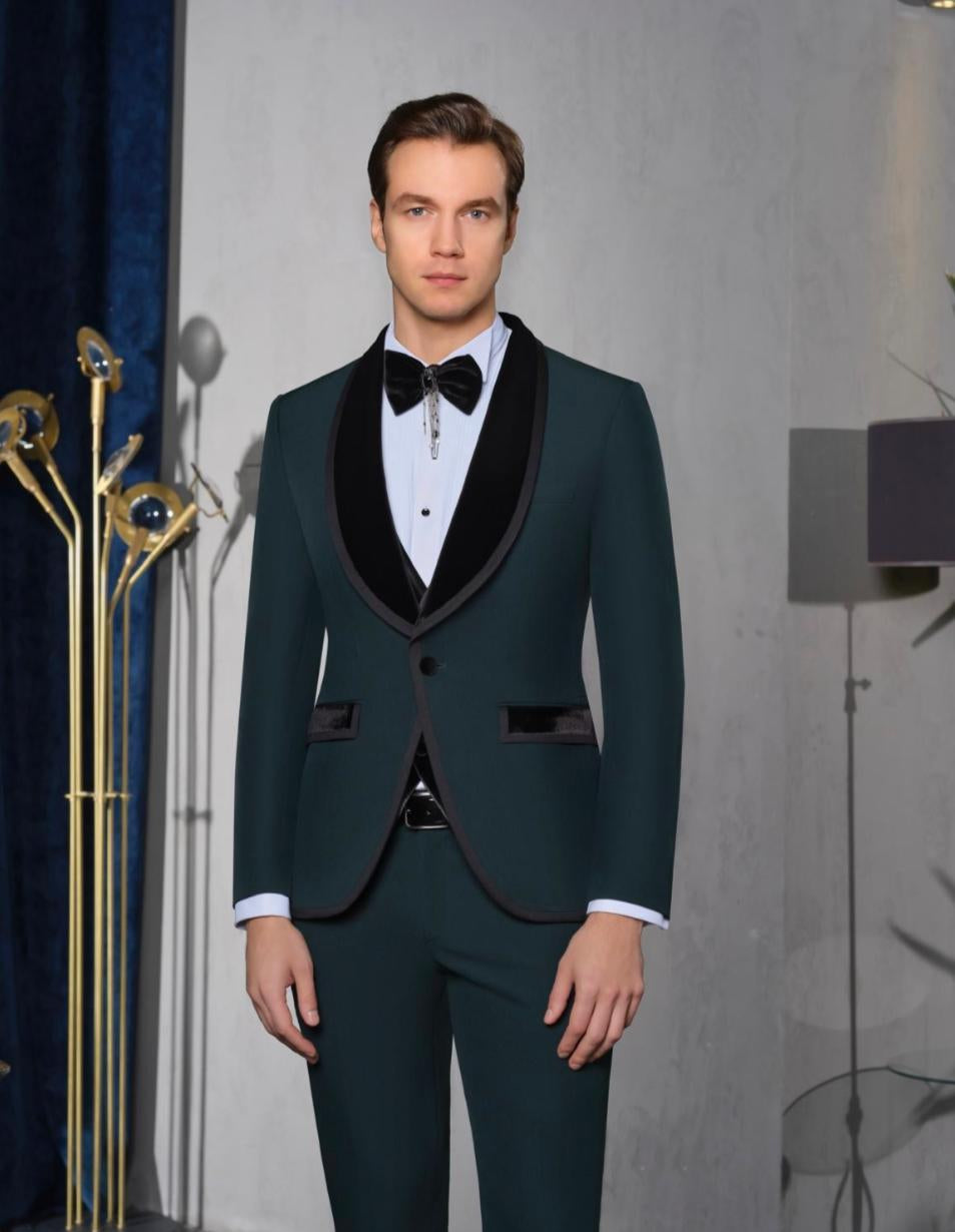 A man wearing a dark teal green tuxedo with black satin lapels, a white dress shirt, and a black bow tie, standing in a studio setting with a gray wall background and decorative gold floor lamps.