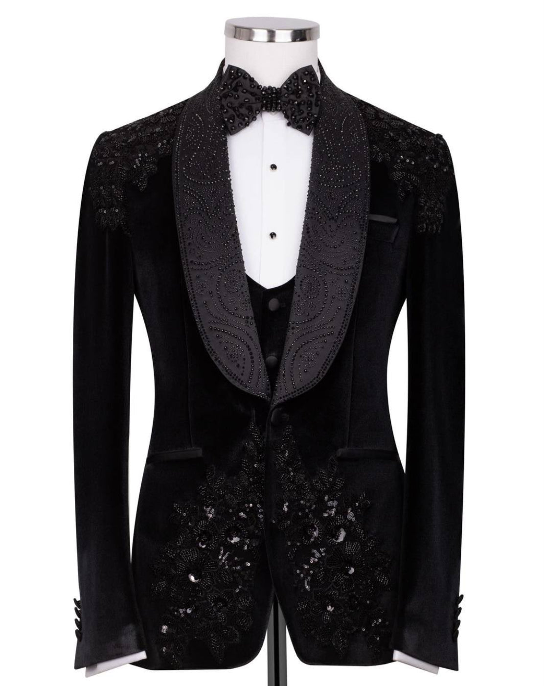 A black velvet tuxedo jacket with ornate beaded and sequined lapels and front panels, displayed on a mannequin with a white dress shirt, black vest, and matching bow tie 