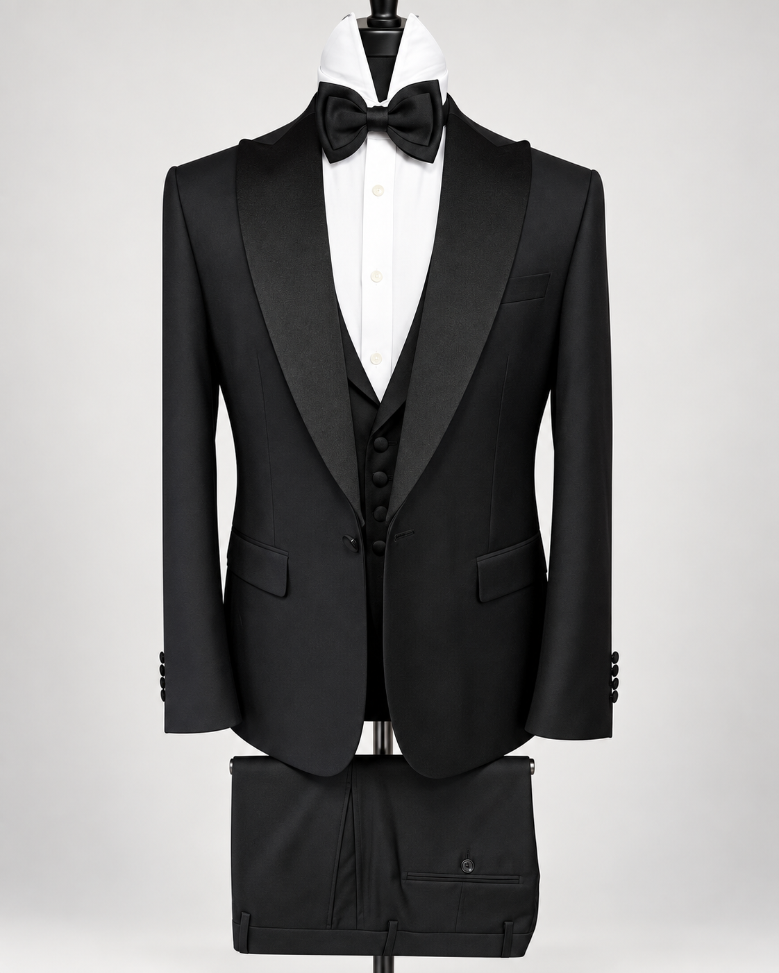 A black tuxedo jacket with satin peak lapels, displayed on a mannequin with a white dress shirt, black bow tie, matching black vest, and black trousers against a light gray background.