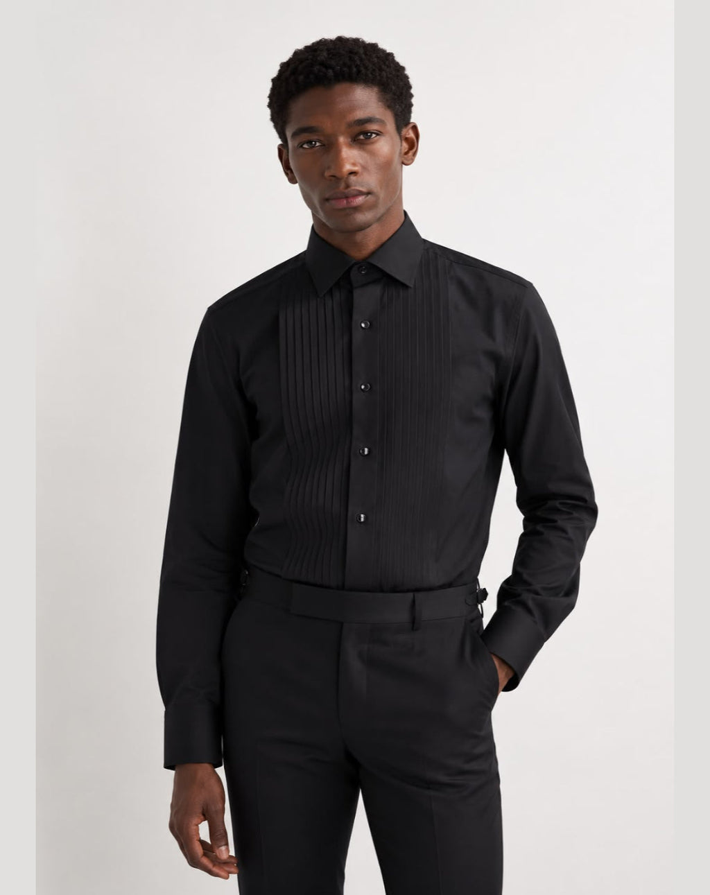 A man wearing a black long-sleeve button-up shirt with vertical pleats on the chest, paired with black trousers, photographed against a plain light gray studio background.