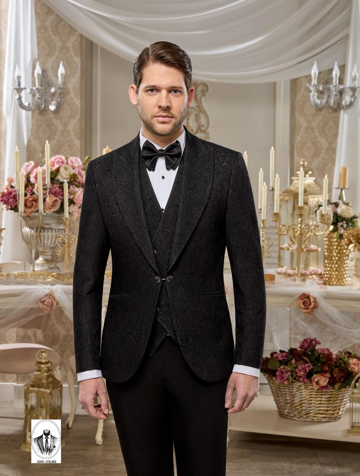 A man wearing a dark charcoal gray three-piece tuxedo with peak lapels, a matching vest, a white dress shirt, and a black bow tie, standing in an elegant, decorated room with draped white fabric, candelabras, and floral arrangements.
