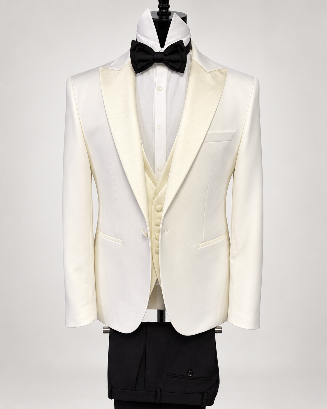 An ivory white tuxedo jacket with satin shawl lapels, styled on a mannequin with a cream vest, white dress shirt, black bow tie, and black trousers against a light gray background.