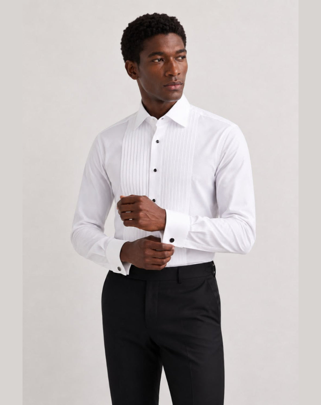 A man wearing a white long-sleeve pleated dress shirt and black trousers, standing against a light gray studio background.