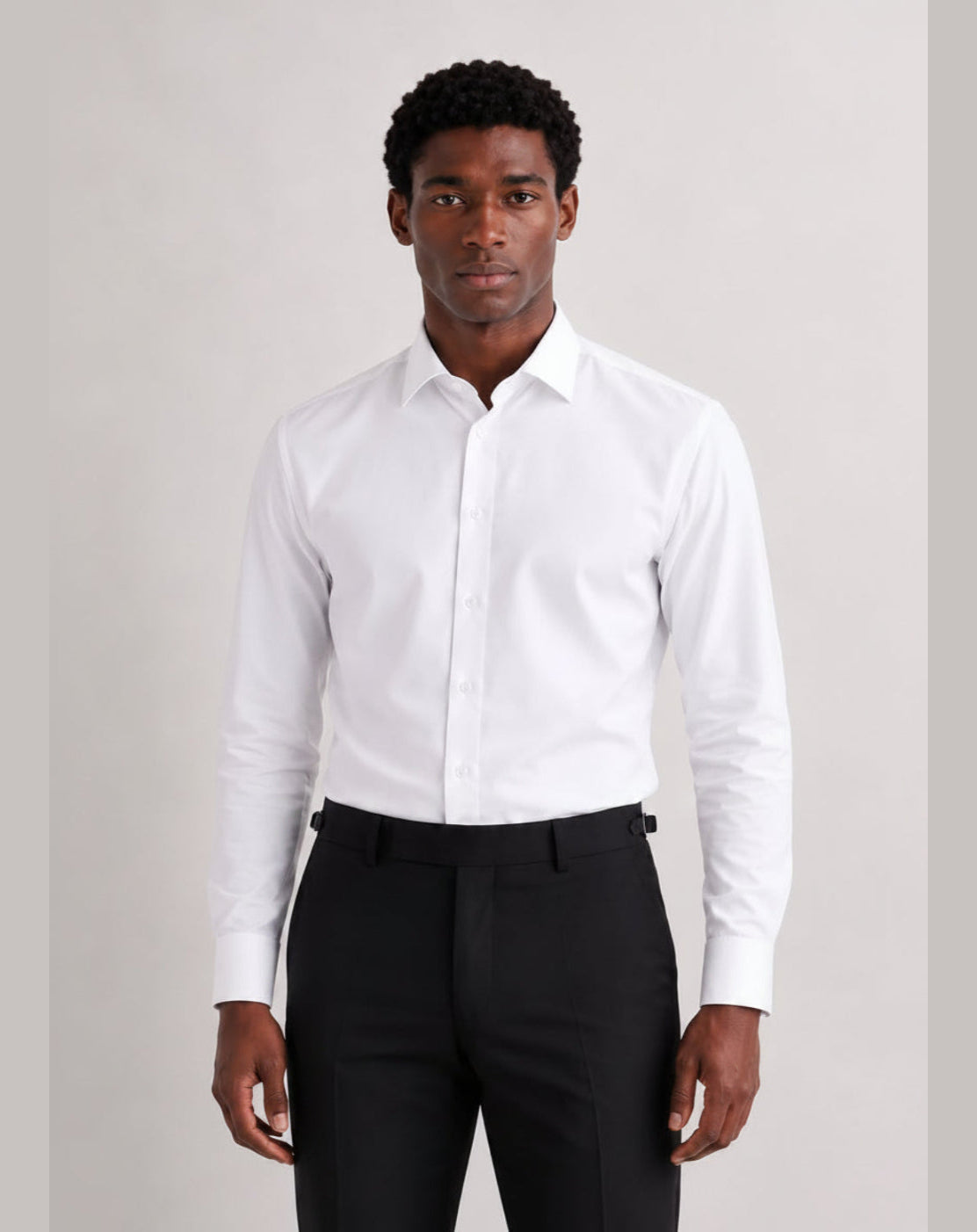 A man wearing a white long-sleeve button-down dress shirt and black trousers, standing against a light gray studio background.